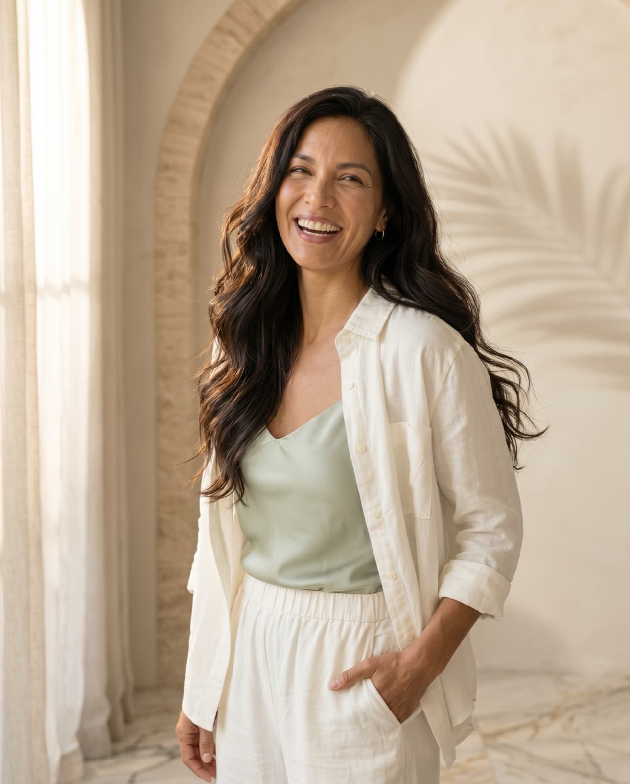 Woman with long, glossy dark hair laughing beside a sunlit arched window, one hand in the pocket of her linen trousers